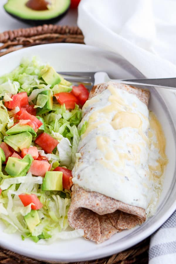 Burrito with white sauce and cheese next to a salad of lettuce, tomatoes, and avocado