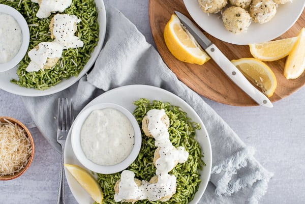 White bowl of green rice and chicken ricotta meatballs covered in a white sauce with lemons