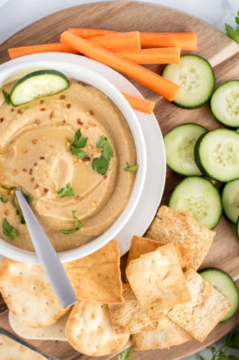 white bowl of hummus with crackers, vegetables, and herbs
