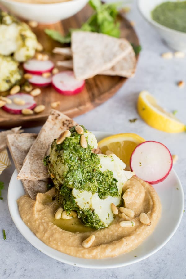 wedge of cauliflower on a plate of hummus with green chimichurri sauce over top