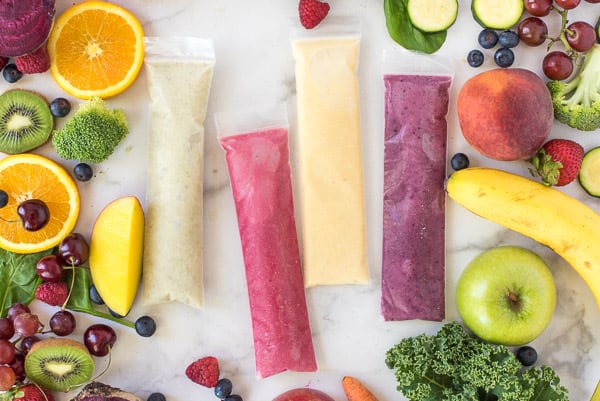 green, pink, yellow, and purple yogurt tubes in a row surrounded by fruits and vegetables