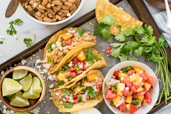 three tacos with chicken and peach salsa next to a bowl of beans and cilantro
