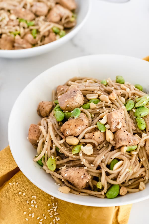 White bowl of noodles, chicken, and edamame with peanuts on top