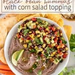 Black bean hummus in a white bowl topped with corn salad, surrounded by chips and avocado