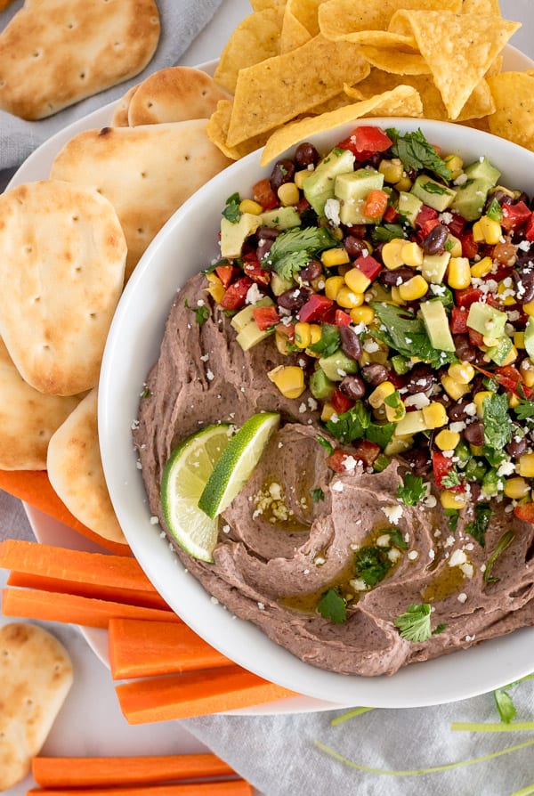 Black bean hummus in a white bowl topped with corn salad, surrounded by chips and avocado