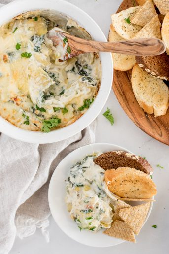 spinach artichoke dip in a white bowl being scooped with a wooden spoon