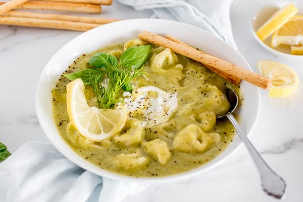 zucchini tortellini soup in a white bowl garnished with lemon and basil
